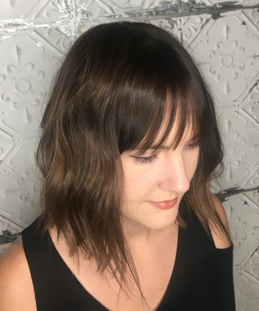 A woman with a long, texturized brunette long bob featuring choppy layers and thin, piecey bangs.