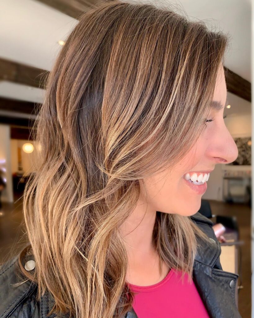 A side profile of a woman smiling with a long, wavy brunette bob featuring heavy honey blonde balayage highlights.