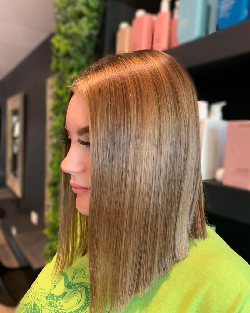 A profile view of a woman with a very straight and shiny honey-toned long bob featuring blunt ends.