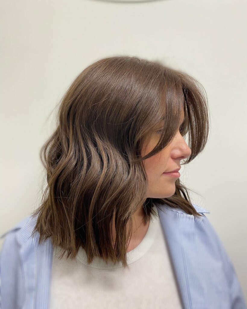 A woman in profile showing a collarbone-length brunette lob with soft waves and light, wispy bangs.