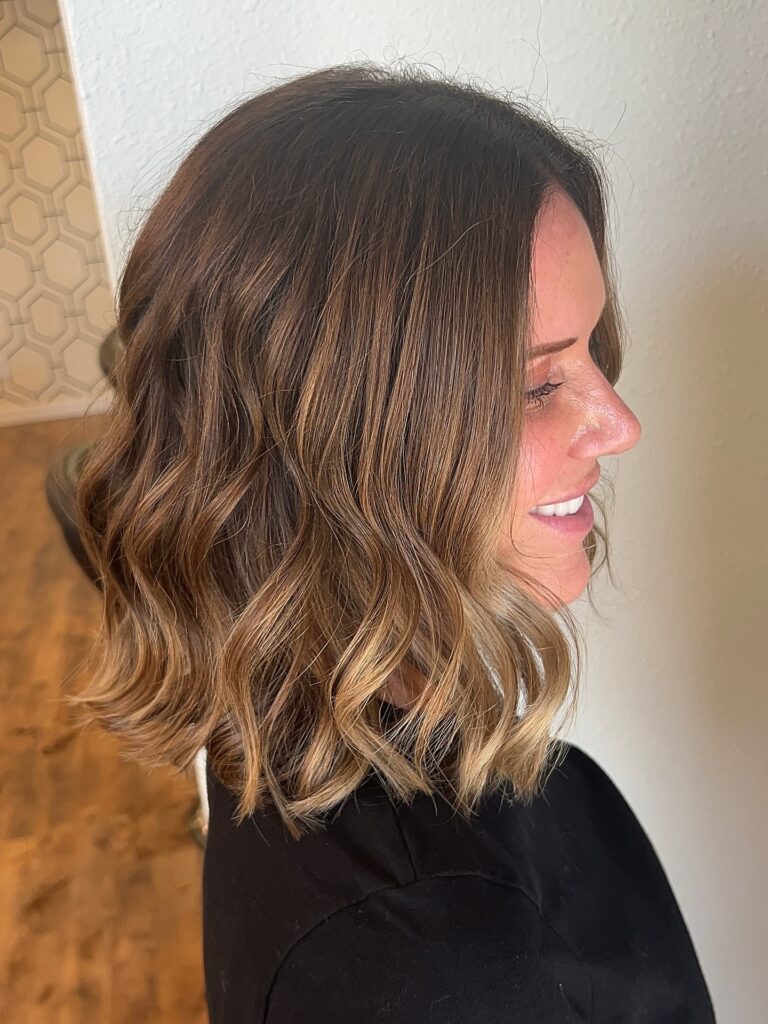 A side profile of a woman smiling with a wavy brunette long bob that has golden toffee highlights.