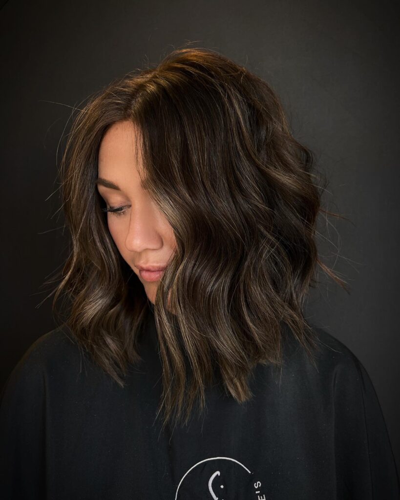 A woman with a textured, ashy brunette long bob styled in loose, undone waves with a middle part.