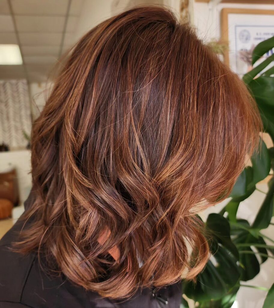 A side view of a woman with a collarbone-length auburn-brunette lob featuring choppy layers and soft, tousled curls.