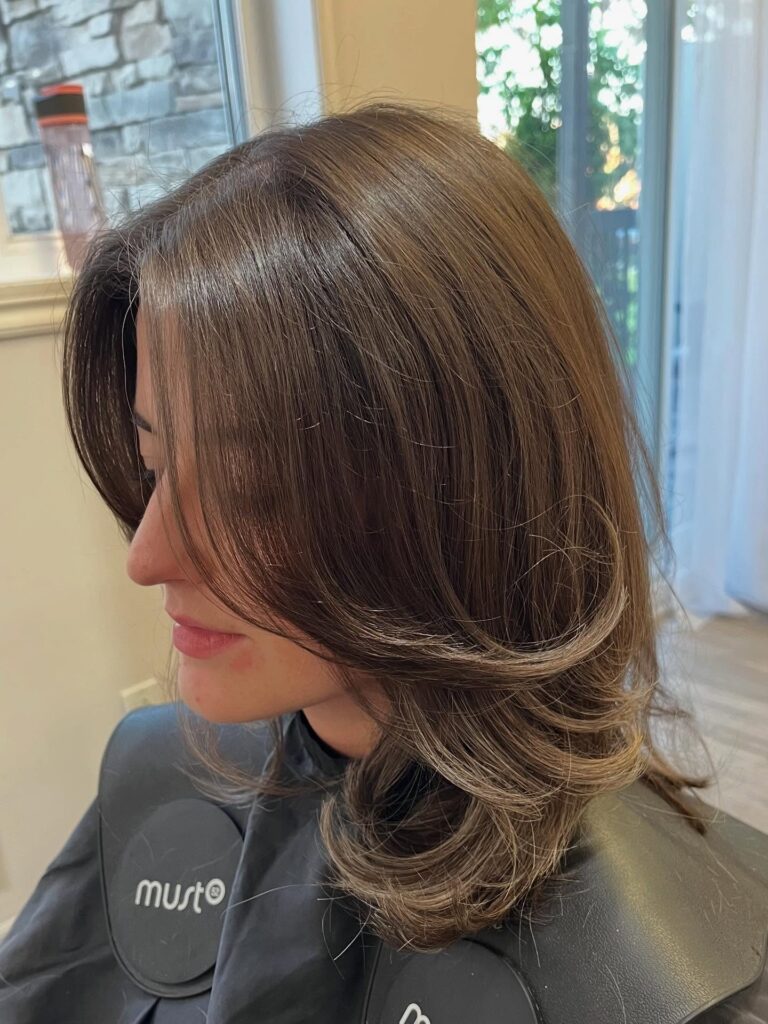 A side profile of a woman with a bouncy brunette long bob featuring dramatic outward-flipped layers and curtain bangs.