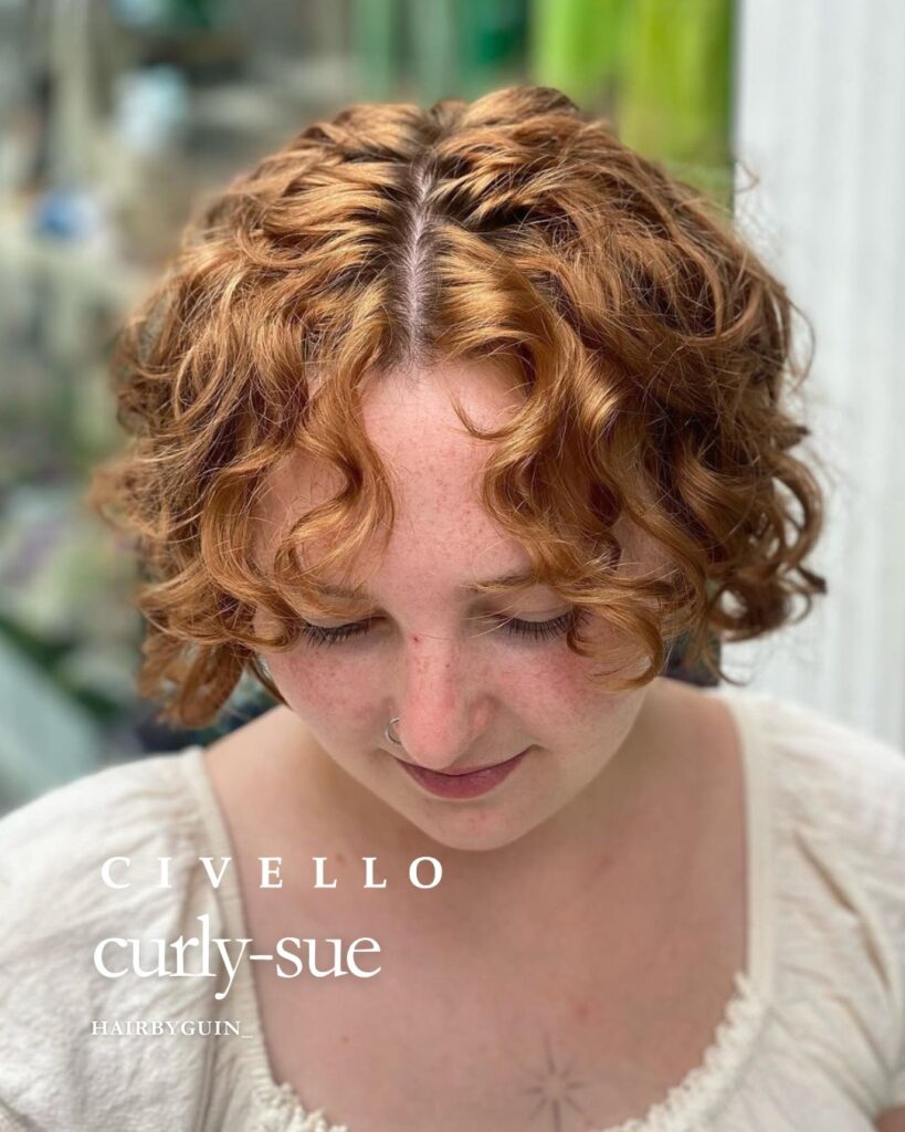 A woman with a short, ear-length, copper, curly bob that is parted down the middle with soft, wavy bangs.