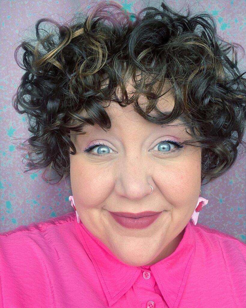 A woman with a short, voluminous curly black bob featuring subtle gold highlights and thick curly bangs.