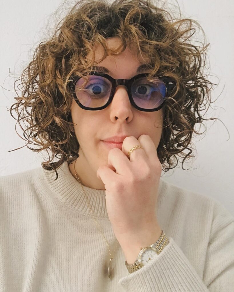 A woman with a honey brown curly bob and matching curly bangs, wearing black, thick-rimmed glasses.