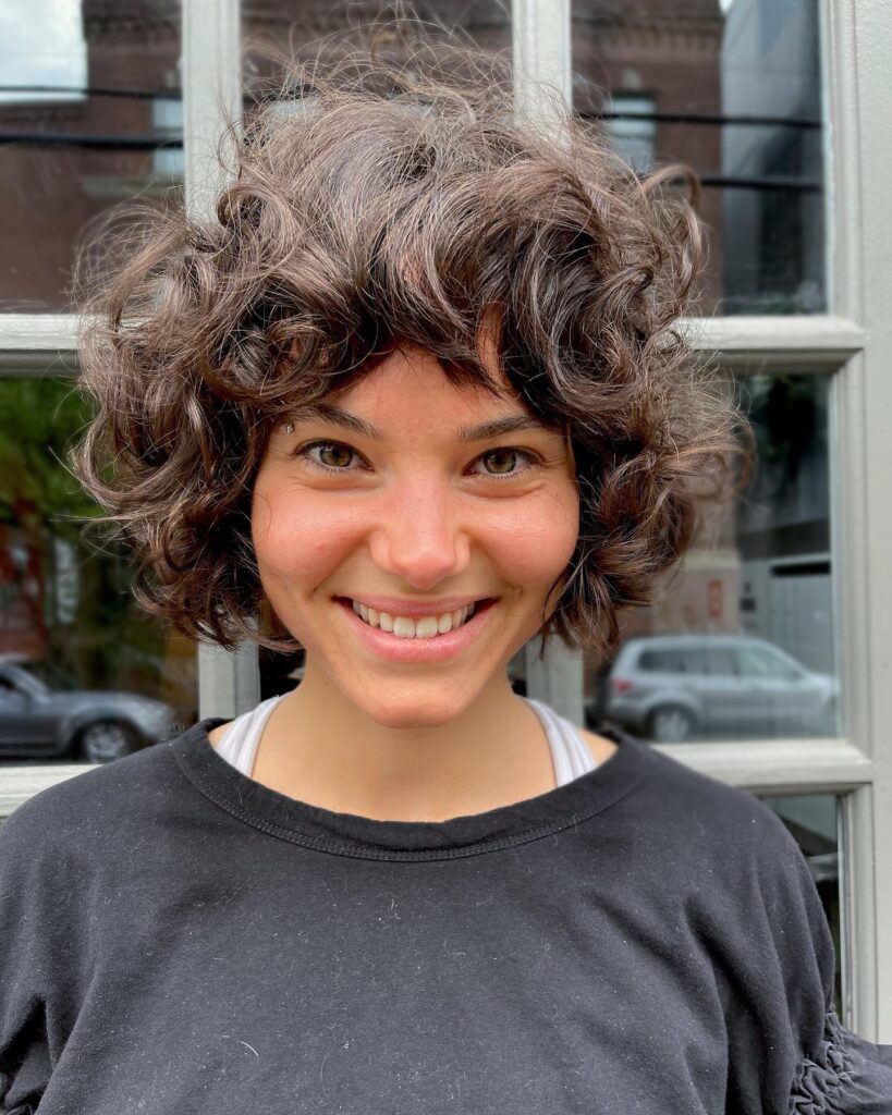 A smiling woman with a chin-length messy brunette bob and piecey wavy bangs.