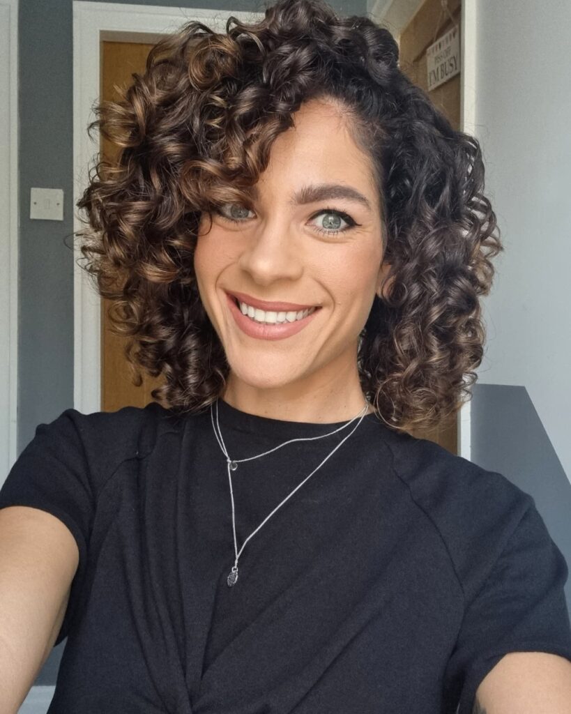 A woman with a shoulder-length curly bob featuring a deep side part, voluminous brunette ringlets, and subtle caramel highlights.