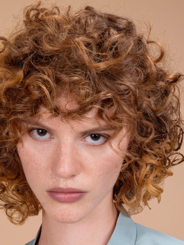 A woman with a neck-length curly auburn bob featuring golden highlights and a thick, tousled fringe.
