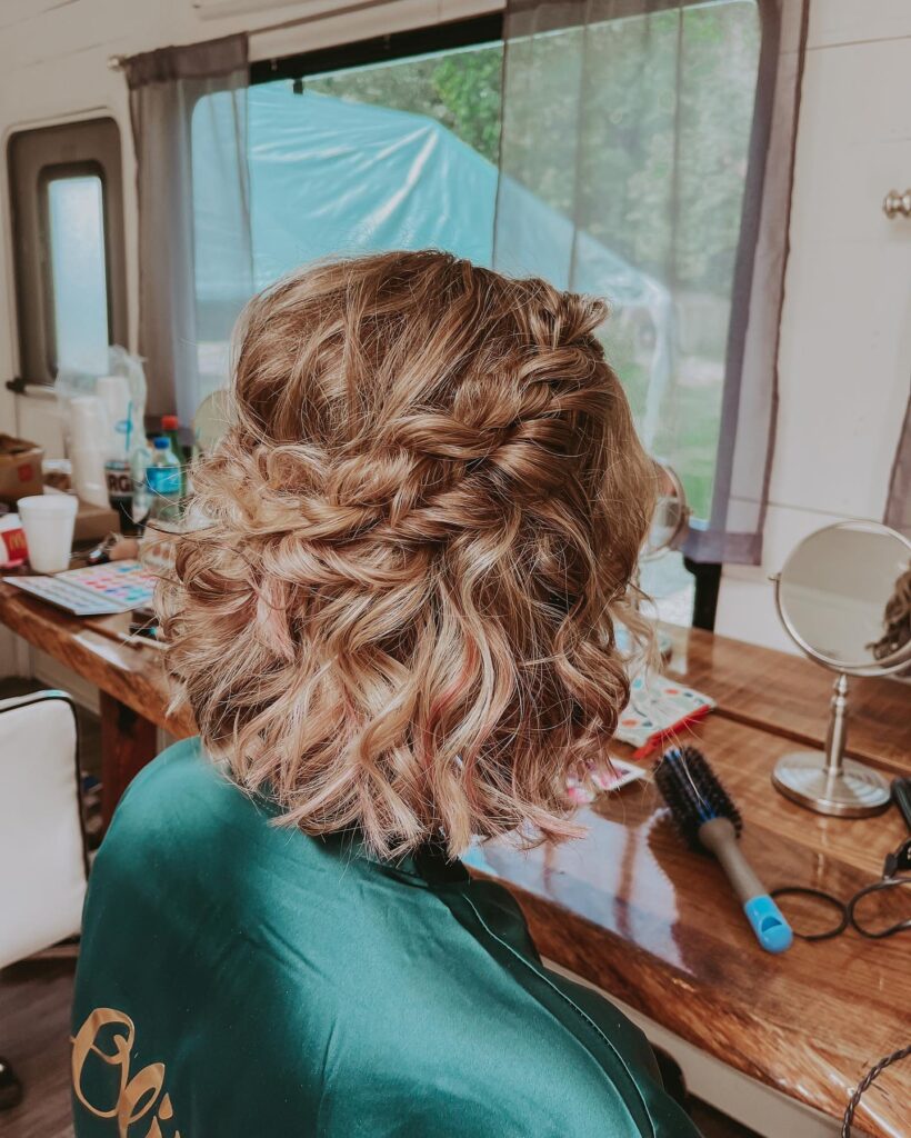 A woman with a shoulder-length curly bronde bob featuring a thick horizontal braid around the back and soft pinkish highlights.