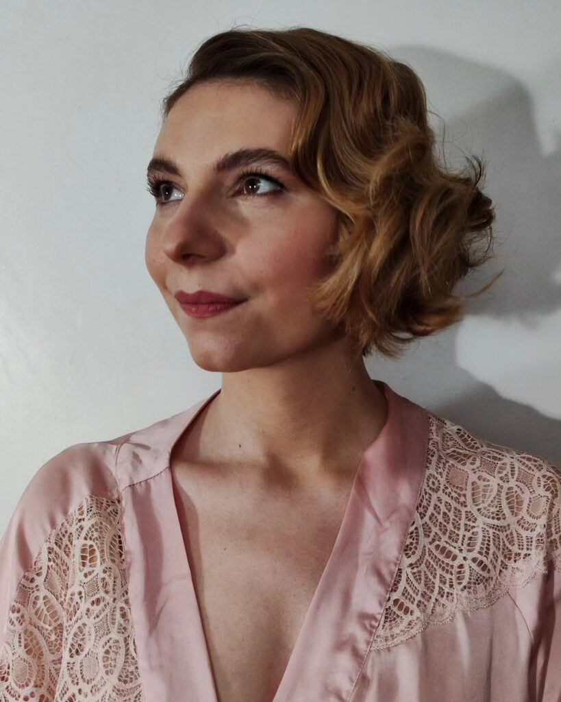 A close-up photograph of a woman with a chic, caramel blonde chin-length bob styled into defined, glossy Old Hollywood waves and a deep side part, looking upward.