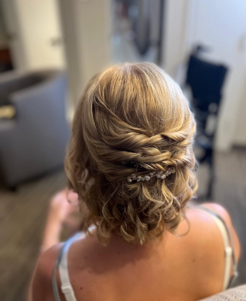 A back view of a woman with a blonde curly bob styled in a twisted half-up look secured with a pearl barrette.