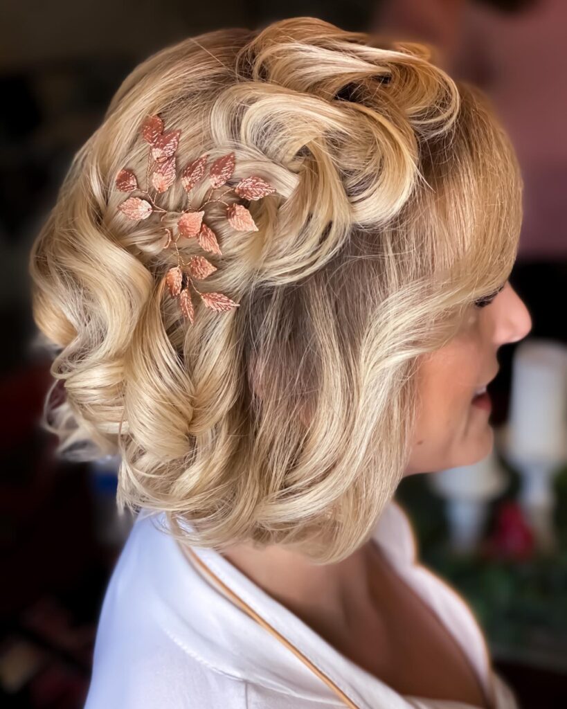 A side view of a woman with a curly vanilla blonde bob featuring a rose gold leaf hair accessory pinned into a side-swept style.