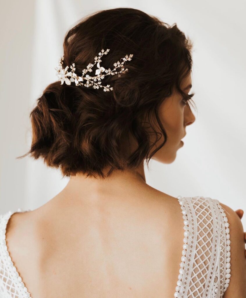 A back view of a woman with a wavy dark brunette bob featuring an ornate crystal and gold floral hair vine.