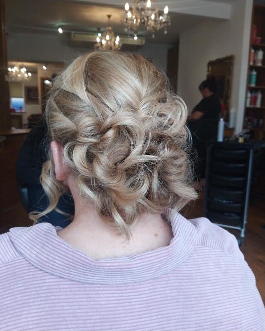 A woman from the back with a curly ash-blonde bob styled into a low pinned updo with intricate swirled layers.