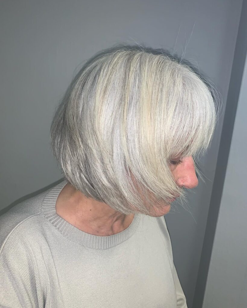 A side profile of a woman over sixty with a silver, choppy bob featuring wispy bangs and soft, textured layers.