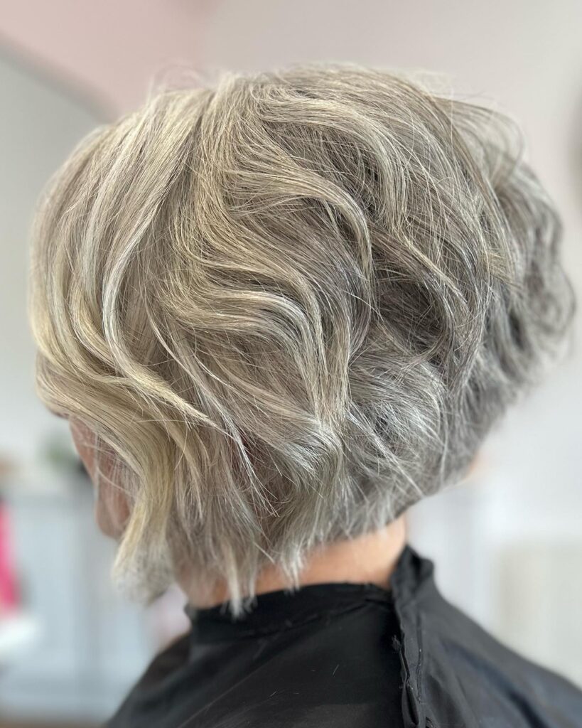 A woman's hair styled in a stacked choppy bob with wavy silver and ash-blonde layers is viewed from the back and side.