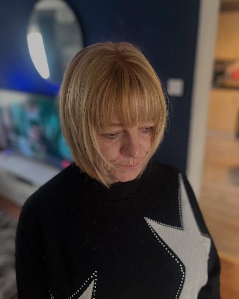 A woman over sixty with a golden honey blonde choppy bob featuring full wispy bangs and chin-length layered ends.