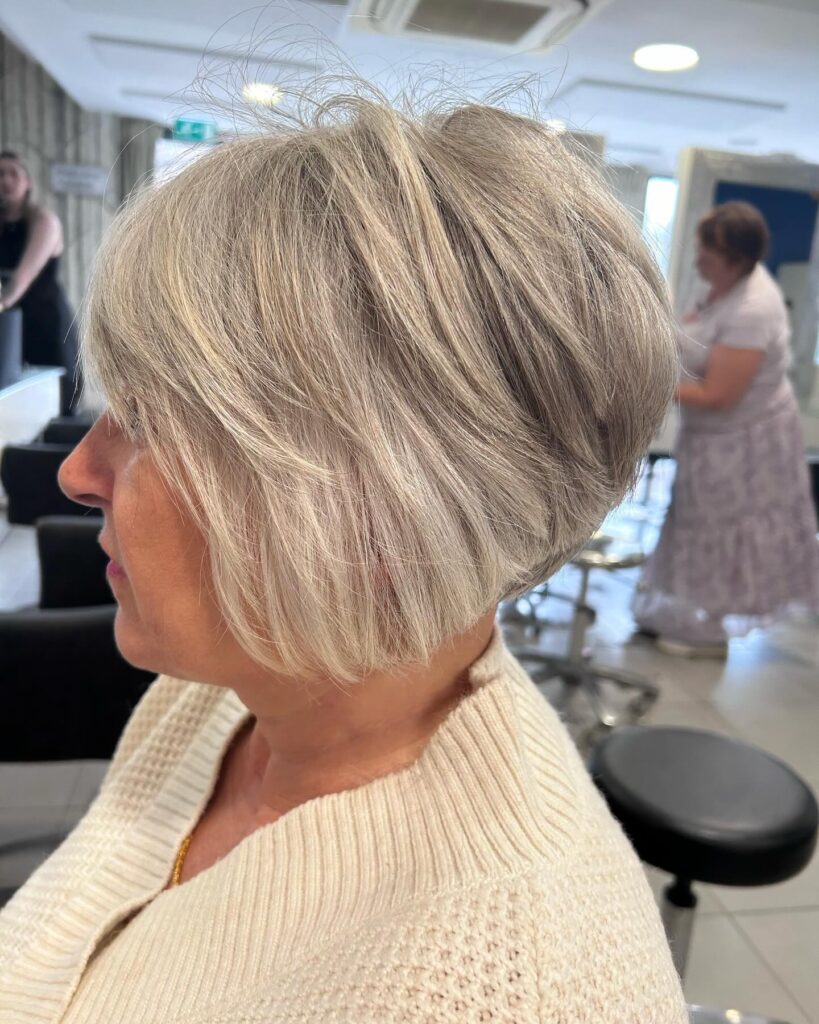 A side profile of a woman over sixty with an angled icy ash-blonde bob featuring stacked choppy layers and a side-swept fringe.