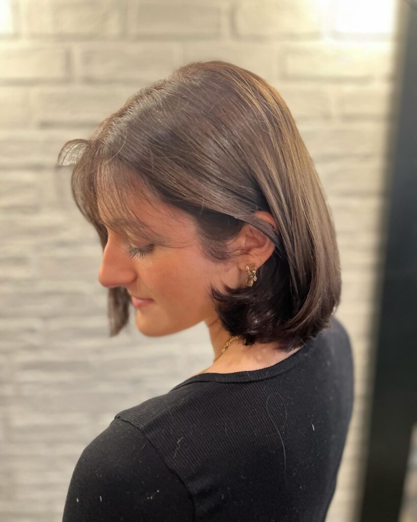 A side profile view of a woman with a straight, neck-length chestnut brunette bob featuring curled-under ends and a wispy fringe.