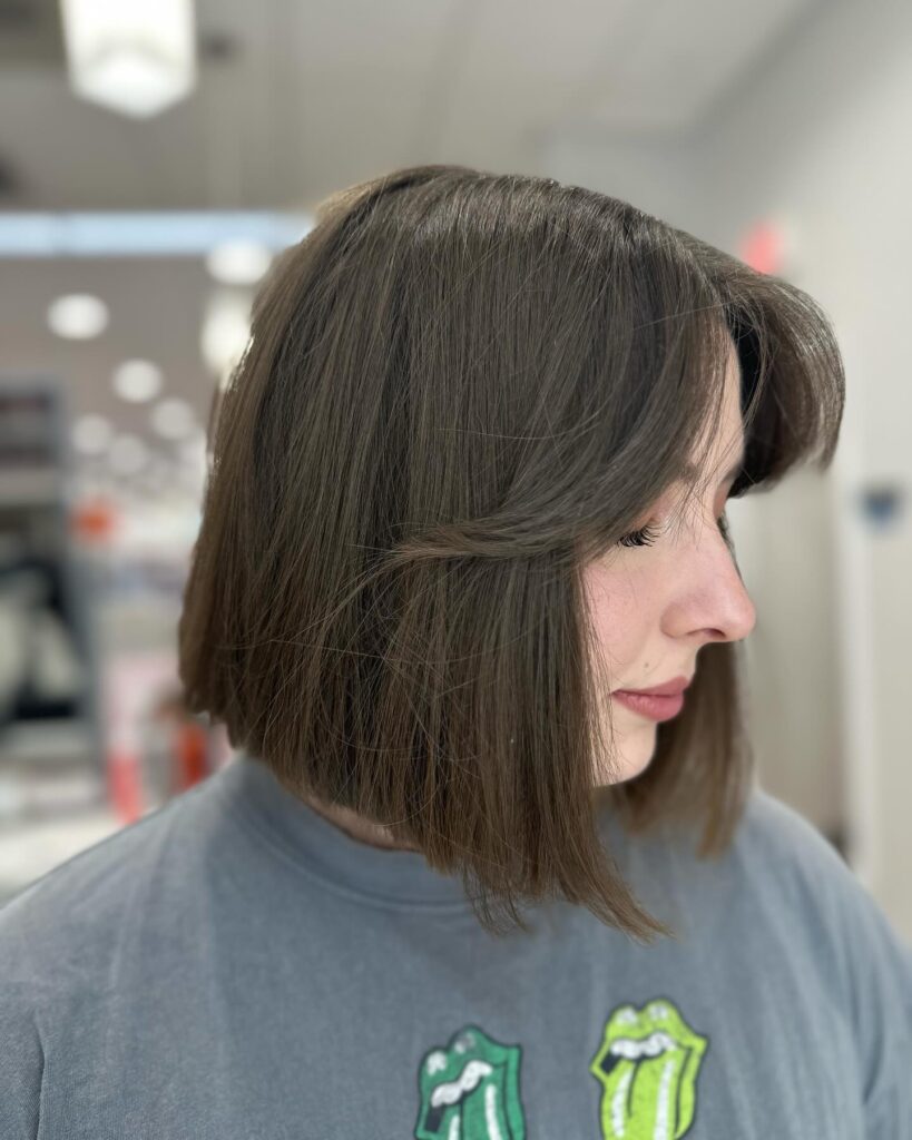 A side profile view of a woman showcasing a straight, neck-length ash brunette A-line bob with soft curtain bangs.