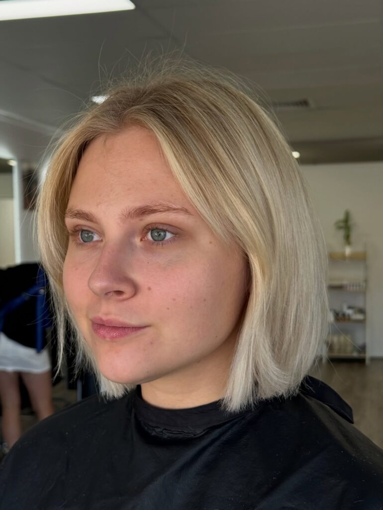 A front view of a woman with a straight, chin-length blonde blunt bob featuring subtly texturized ends and a middle part.
