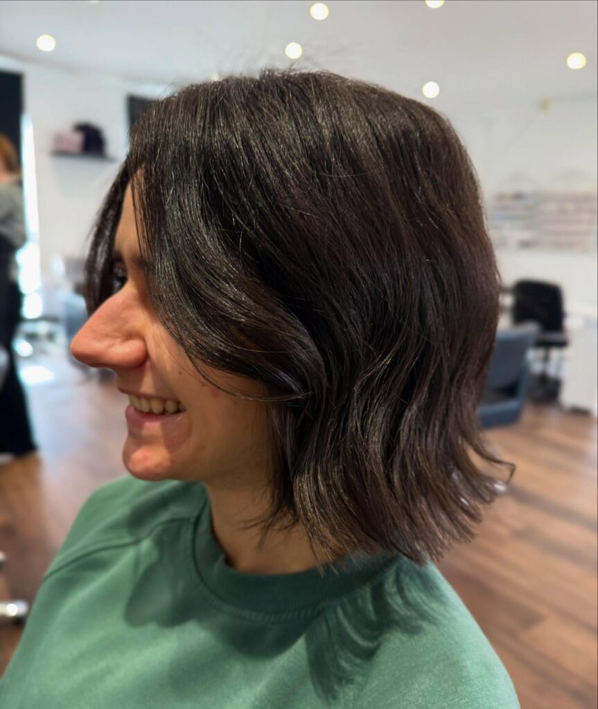 A smiling woman in profile view showcasing a textured neck-length dark brown choppy bob with loose waves and subtly flared ends.