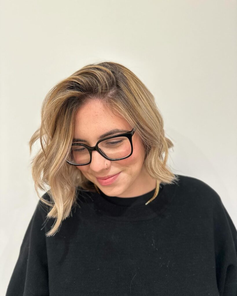 A woman with glasses smiling and showing off a textured blonde choppy bob with a deep side part and loose waves.