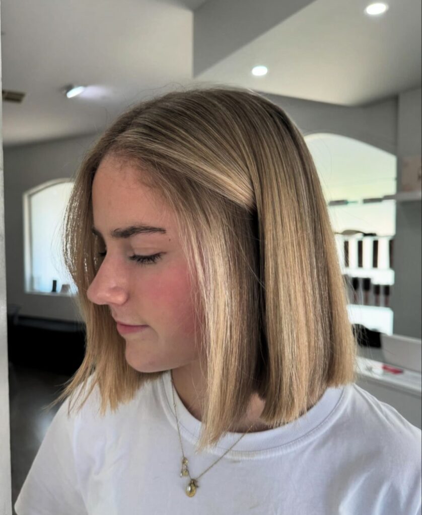 A side view of a young woman with a straight, shoulder-length blunt blonde lob cut with a subtle center part.