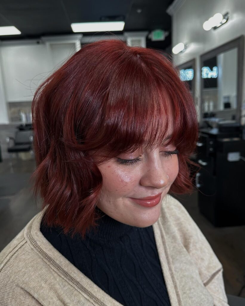A woman smiles showcasing a chin-length textured auburn red bob hairstyle with a full curtain fringe and loose waves.