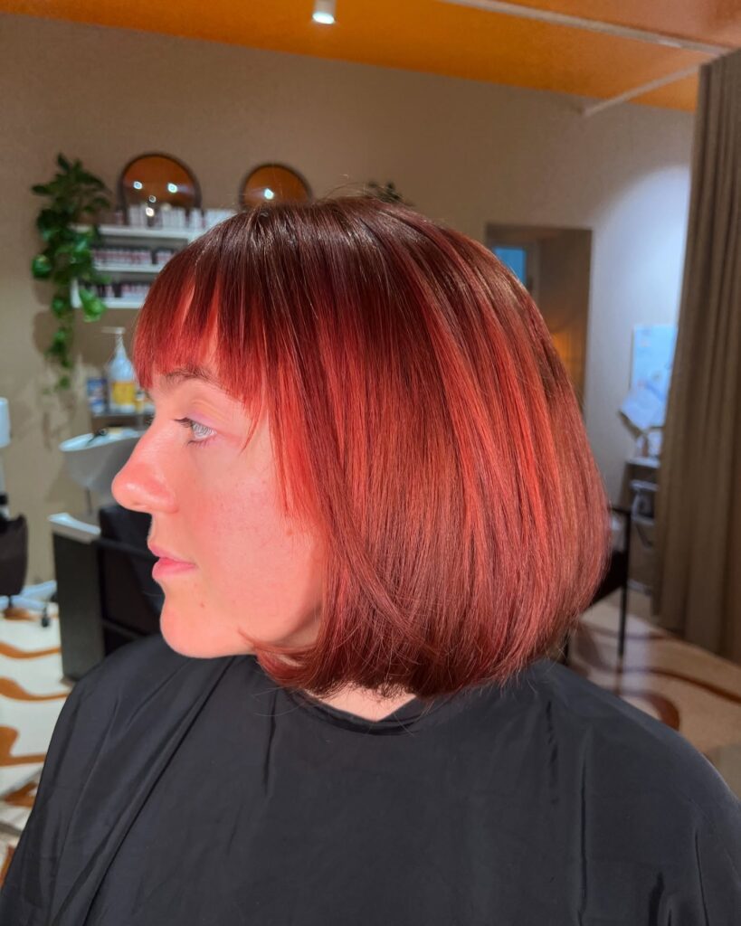 A side profile view of a woman sporting a sleek chin length red bob with copper highlights and full wispy bangs.