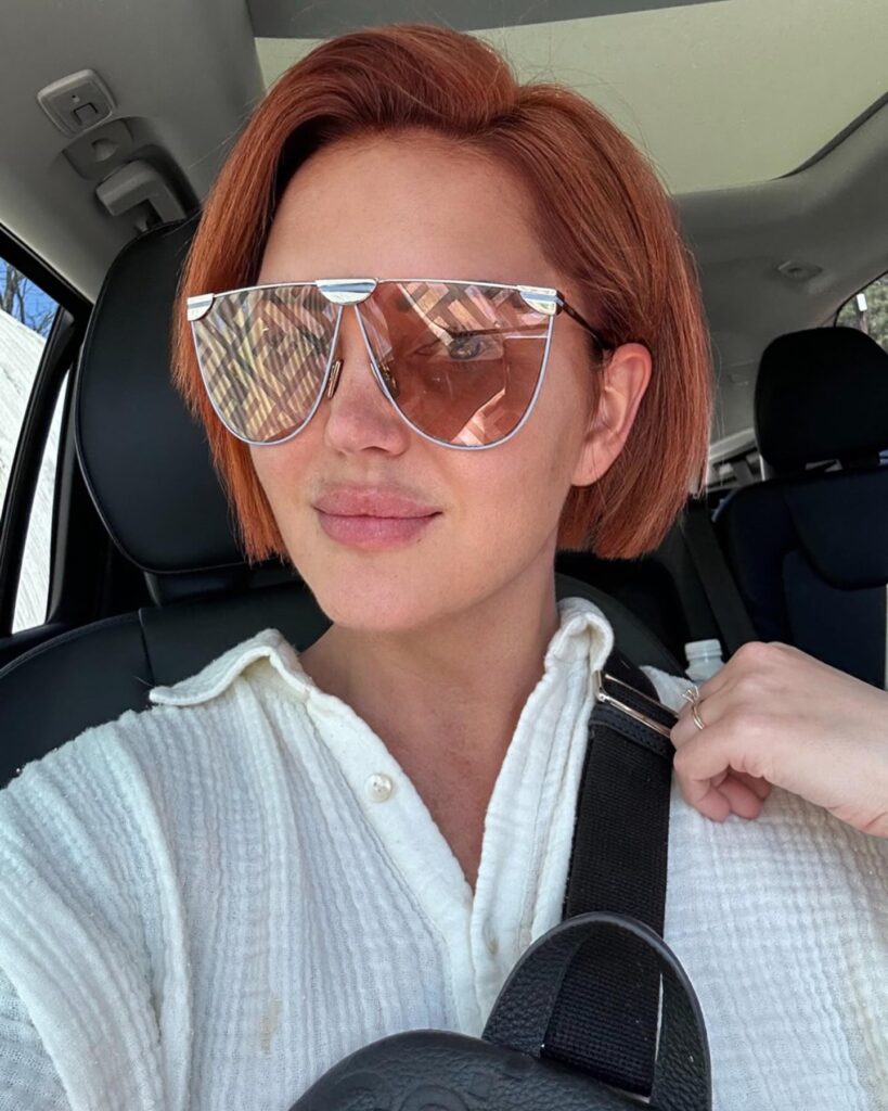 A woman wearing sunglasses in a car showcases a straight, chin-length apricot copper bob hairstyle with a deep side part.