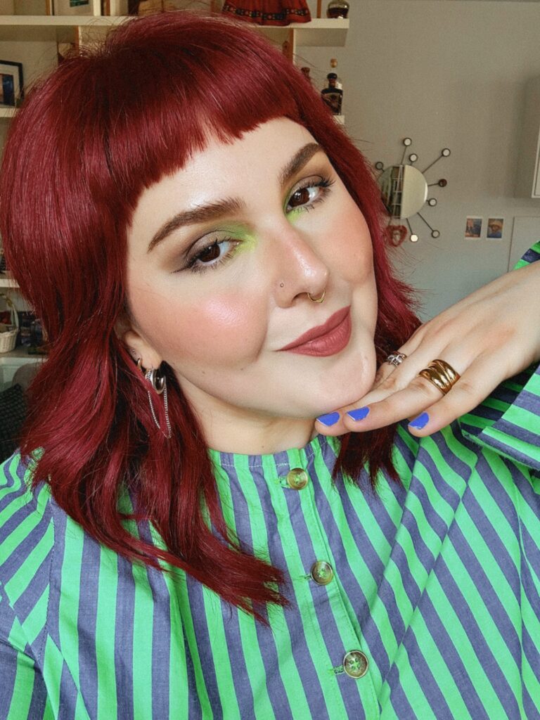 A woman with a textured, neck-length crimson red choppy bob featuring short micro bangs smiling at the camera.