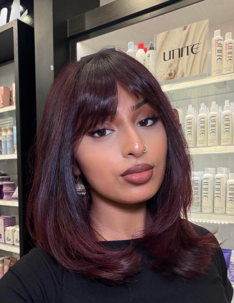 A woman with a smooth, shoulder-length deep plum red long bob featuring soft curtain bangs looking directly at the camera.