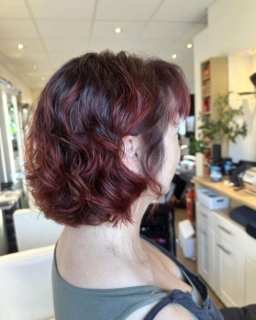 A side profile of a woman showcasing a neck-length curly mahogany bob hairstyle with subtle cherry red highlights.