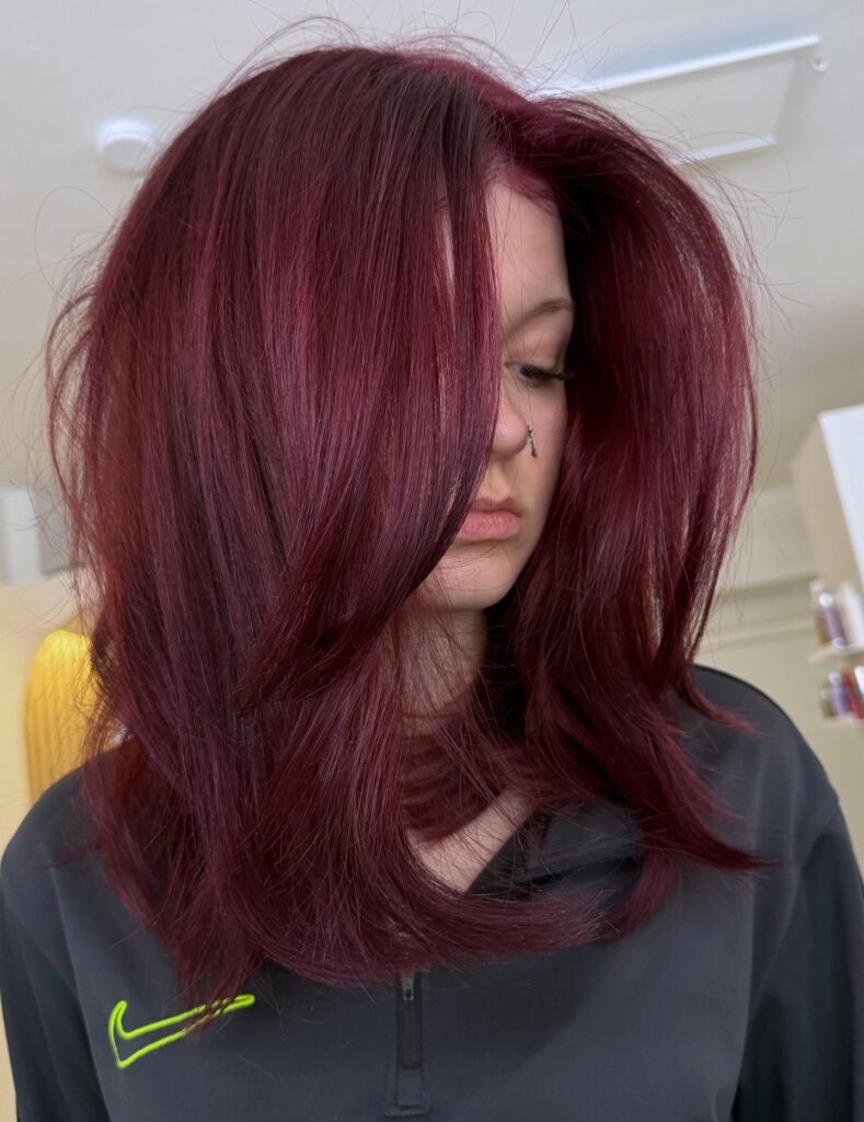 A woman with a voluminous, shoulder length deep burgundy red long bob featuring heavy sweeping layers over her face.