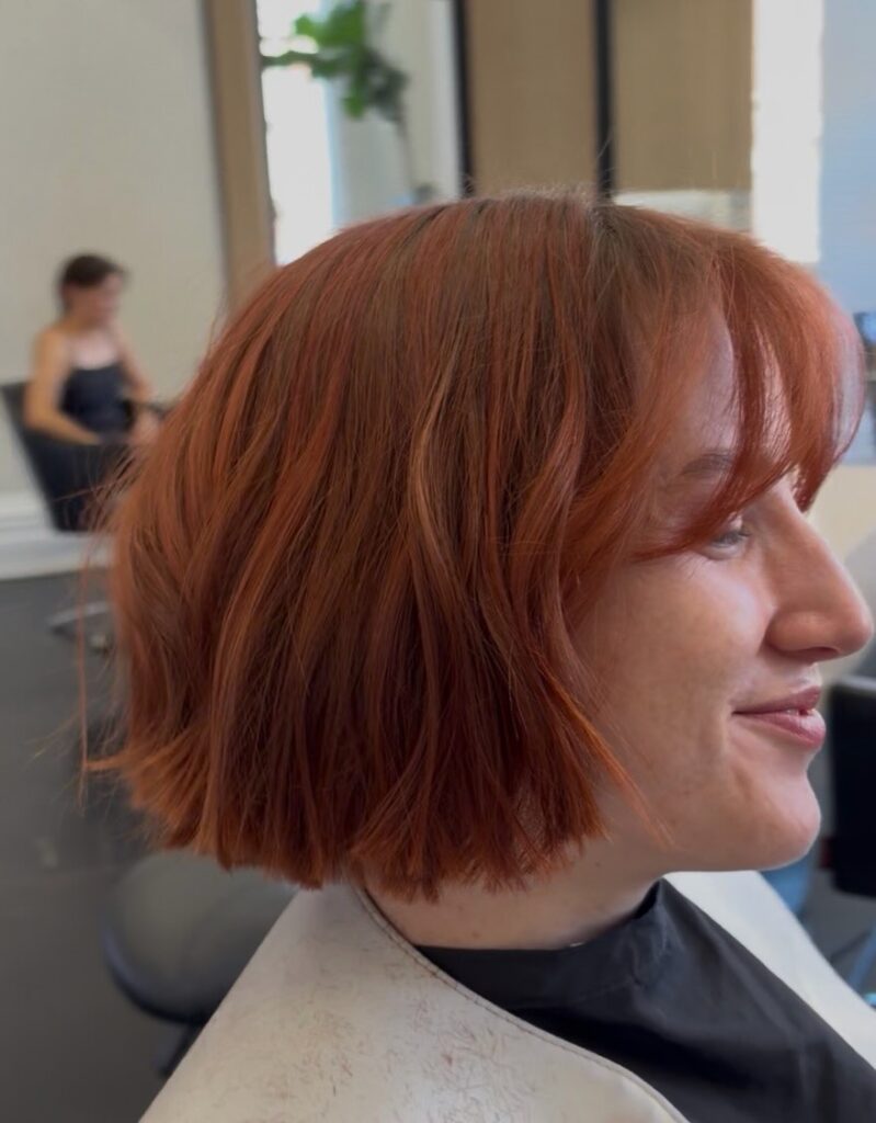 A close-up shot of a woman displaying a textured, chin-length copper red bob with a long, wispy fringe.