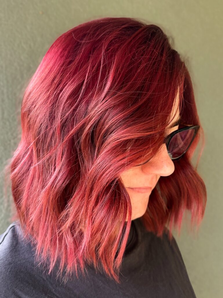A woman from a side angle showcases a textured, shoulder-length magenta red long bob hairstyle styled with messy waves.