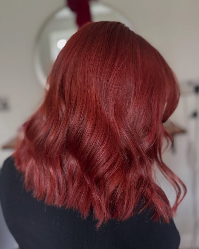 A woman from the back showcases a shoulder-length crimson red long bob hairstyle styled with soft glam waves.
