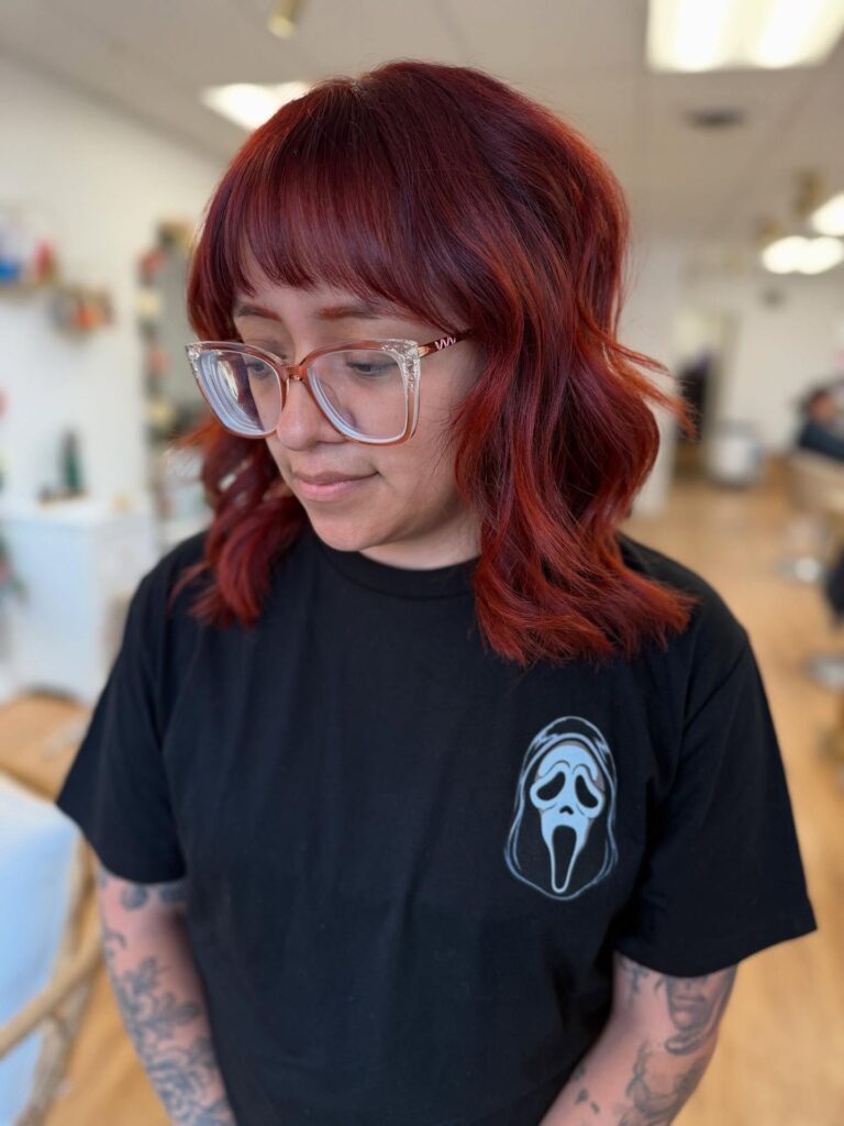 A woman wearing clear glasses showcases a shoulder-length fiery auburn shaggy lob hairstyle with thick blunt bangs and loose waves.