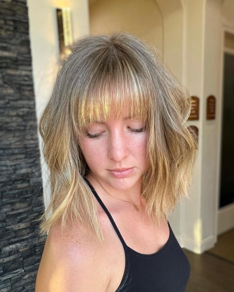 A woman with an ashy blonde long bob featuring wavy texture and a full straight across fringe.