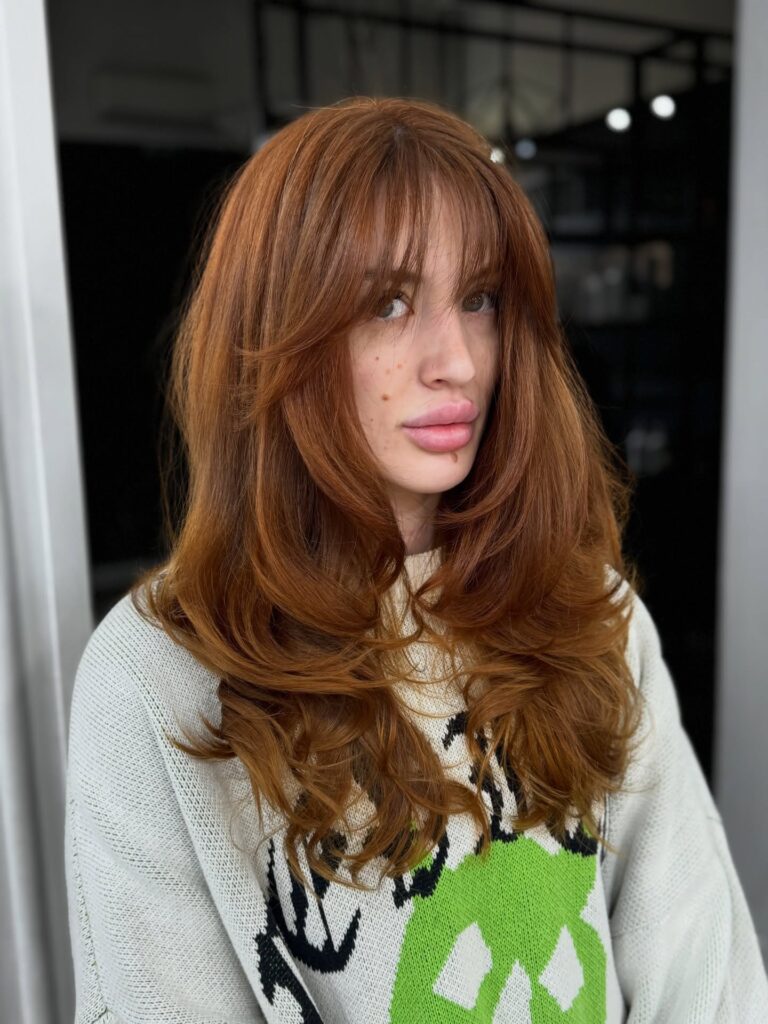 A woman with a voluminous auburn long bob featuring butterfly layers and a wispy face-framing fringe.