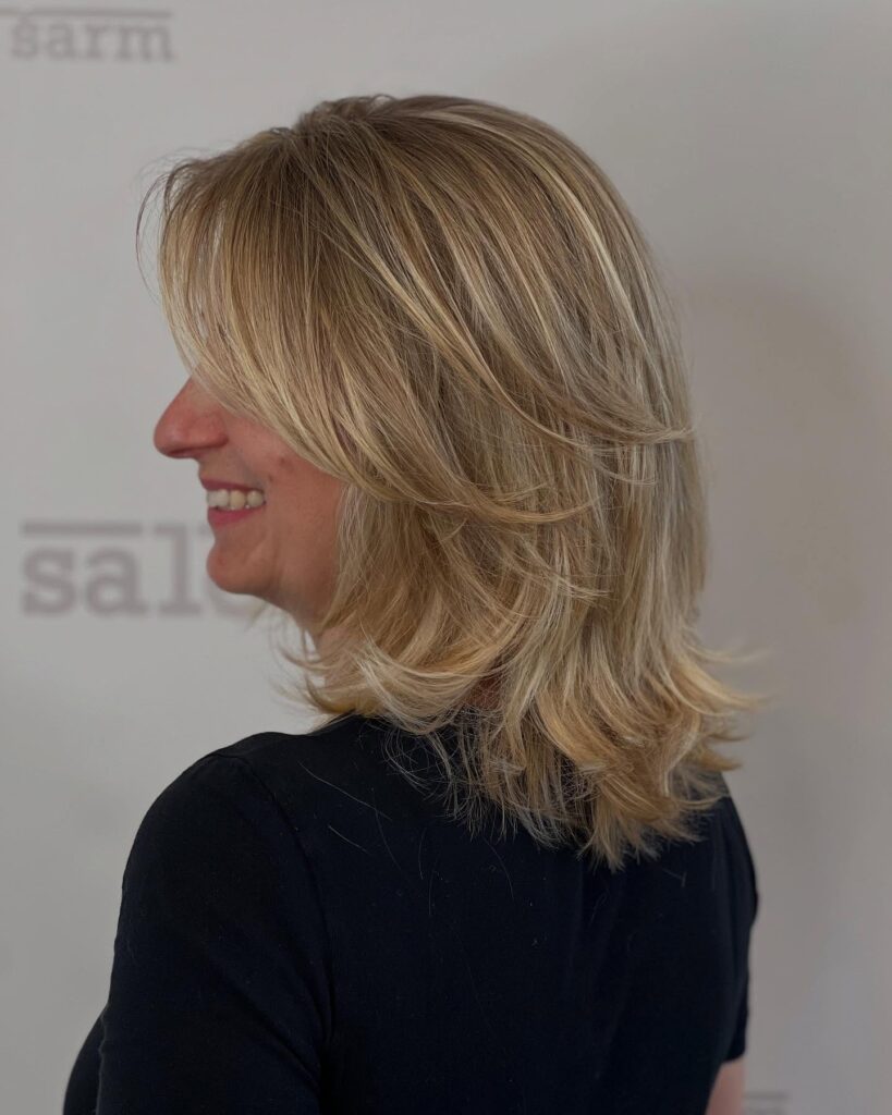 A woman with a shoulder-length layered blonde bob and side-swept bangs showing off the textured volume from a side profile view.