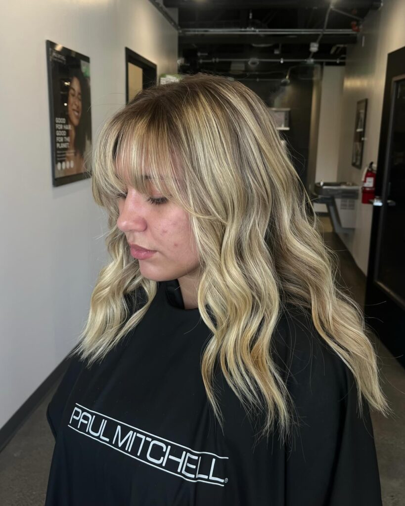 A woman with a shoulder-length rooted golden blonde bob featuring soft waves and parted curtain bangs.