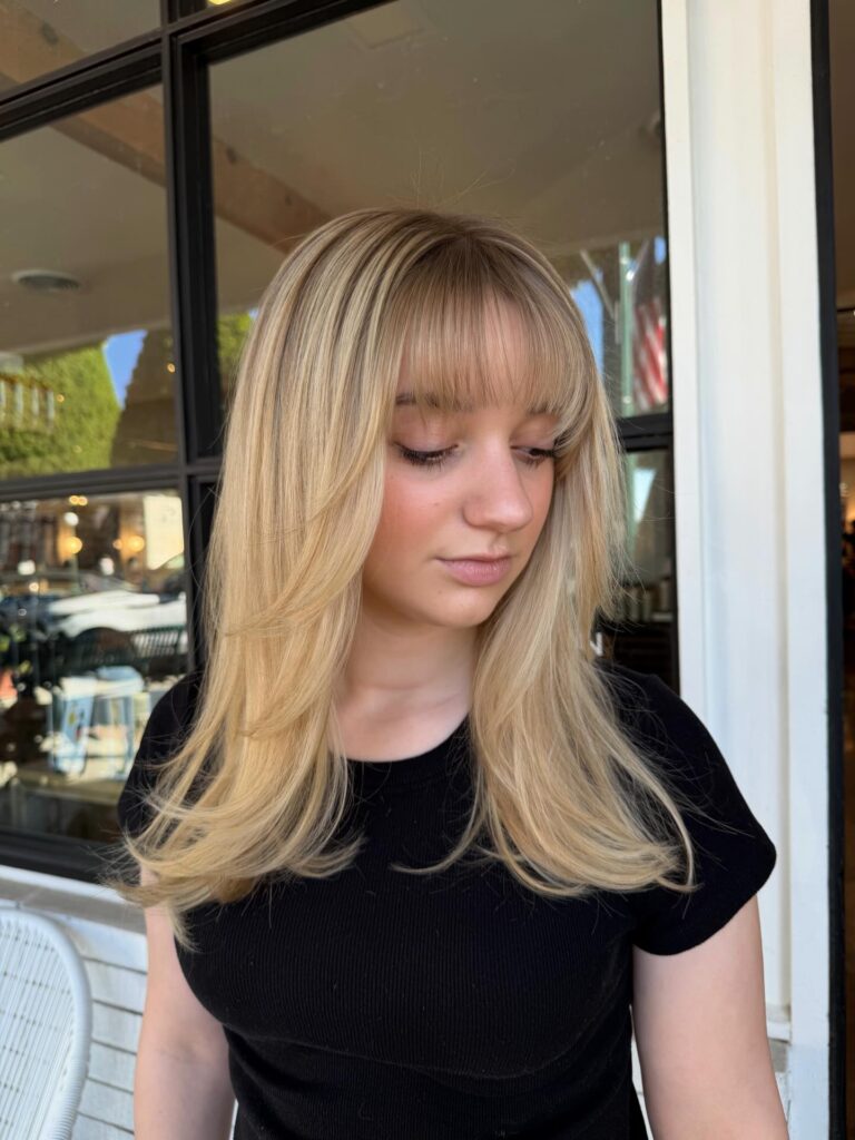 A woman with a polished rooted golden blonde long bob featuring a thick straight full fringe.