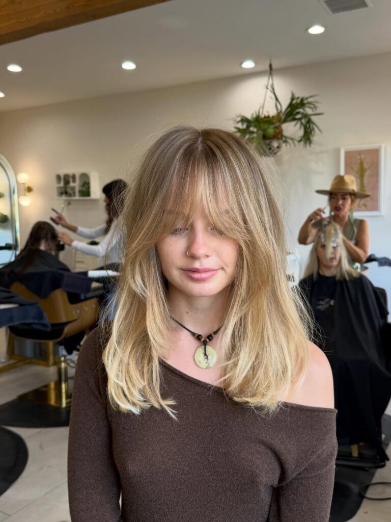 A woman with a shoulder-length rooted golden blonde bob with soft waves and long curtain bangs parting over her eyes.