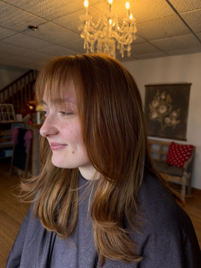 A woman with a warm copper colored long bob featuring wispy bangs and soft flicked out ends.