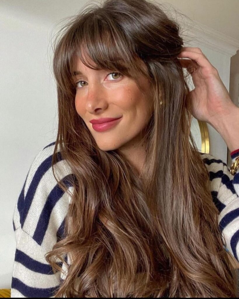 A woman with long chestnut brown hair styled in a long bob with soft waves and wispy front bangs.