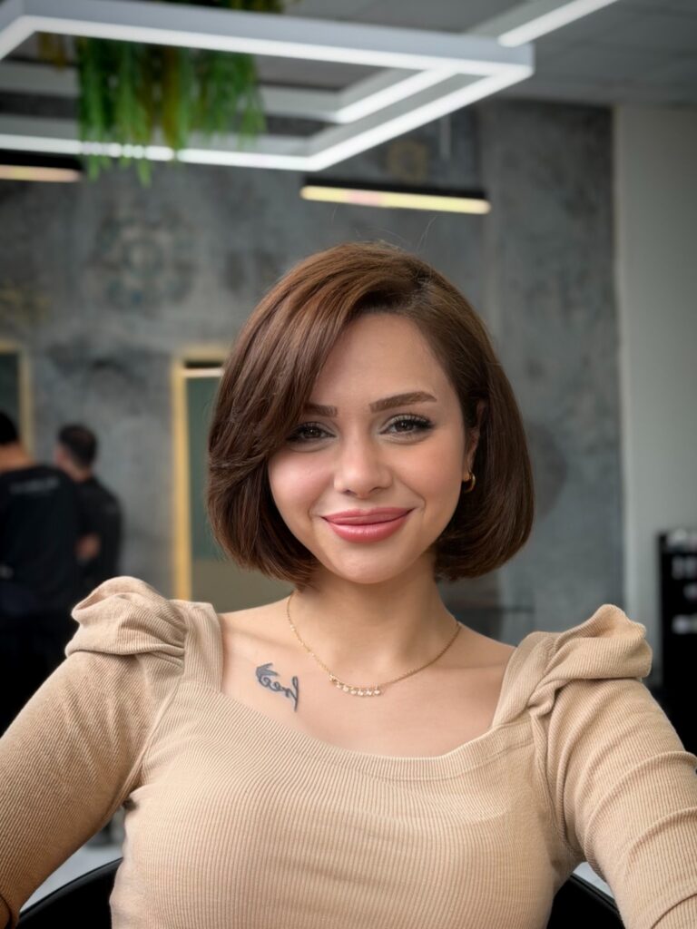 A smiling woman with a smooth, glossy caramel brunette rounded bob featuring a soft side-swept fringe and subtle layers curving inward.